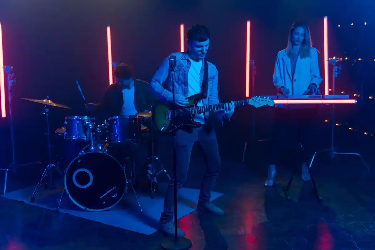 Guitarists and drummer performing on a brightly lit stadium stage with atmospheric lighting