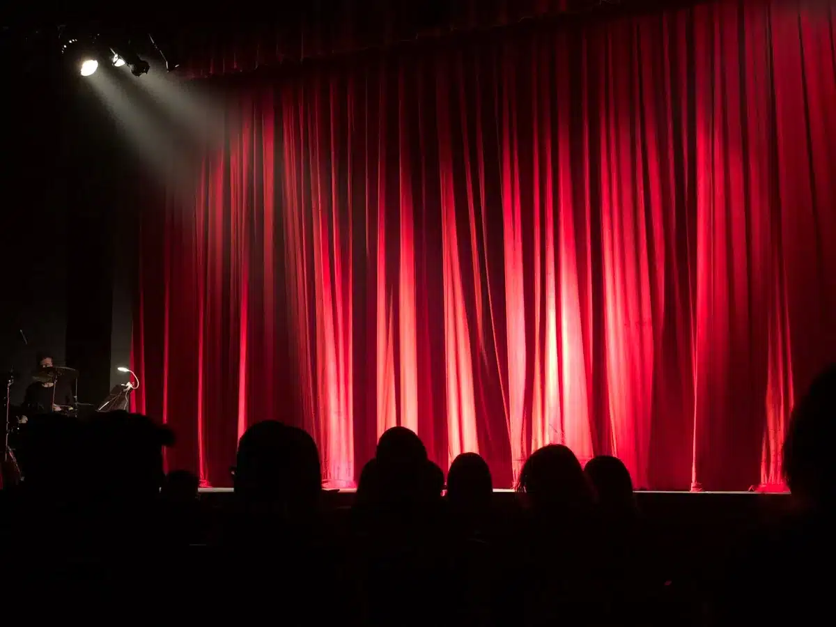 Broadway stage with soft spotlight and audience in respectful silence during a tribute moment