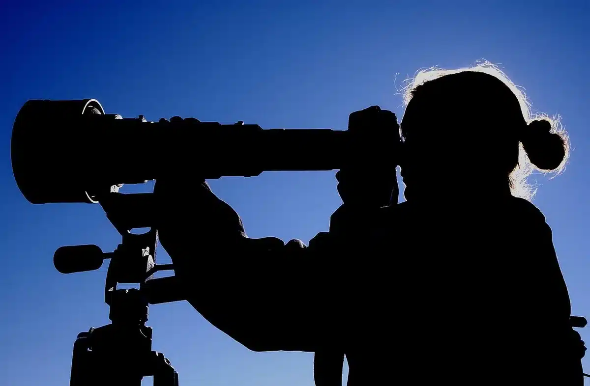 Silhouetted figures and a camera tripod on a rooftop at dusk overlooking a city