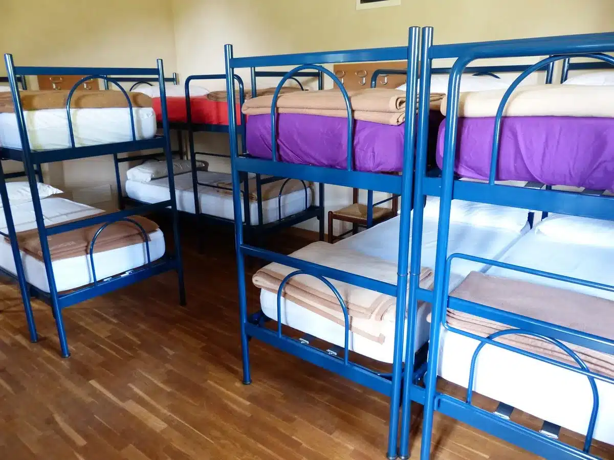 Overcrowded dorm-style sleeping area with bright overhead lights and closely packed beds