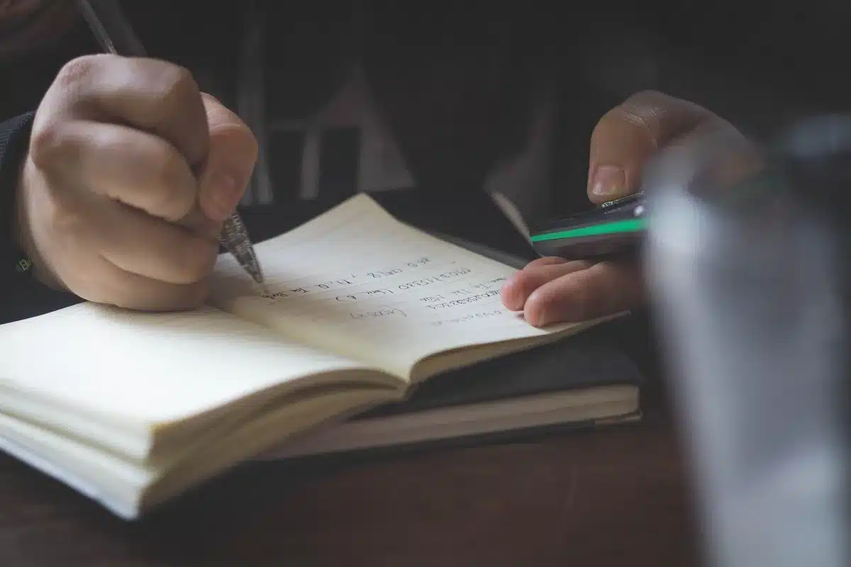 Close-up of hands writing lyrics in a notebook with studio equipment nearby